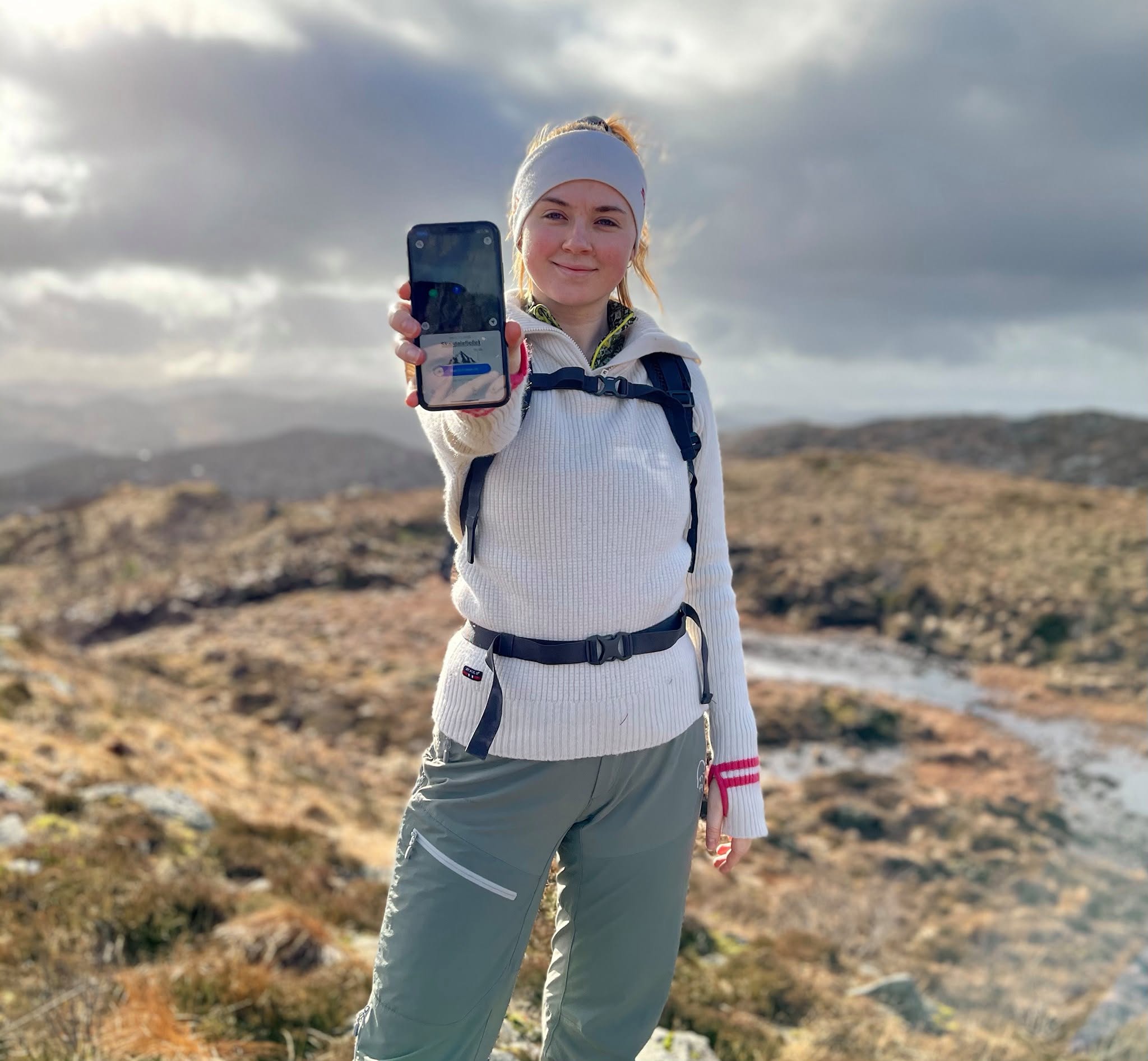 Turgåer på en fjelltopp som holder opp mobilen med Fjelltoppjakten.